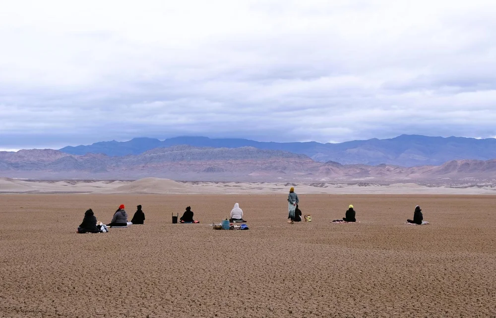 A group sitting apart from one another on a dry desert lakebed, mountains behind